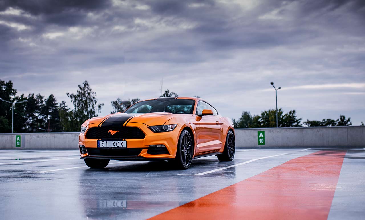 Pomarańczowy Ford Mustang na parkingu na dachu w pochmurny dzień wypożyczalnia Dedicars